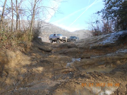 1986 Ford Bronco - Two off-road vehicles, a Ford Bronco and a dark SUV, navigate a very muddy and rutted trail on a sunny day. The Bronco is in the foreground…