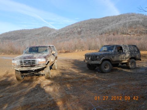 1986 Ford Bronco - Two muddy Ford Broncos are parked on a dirt road with a wooded hillside in the background under a blue sky. The Bronco on the left is dark…