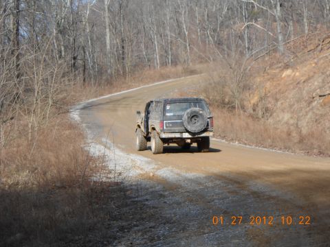 1986 Ford Bronco - Black 1986 Ford Bronco driving on a rural dirt road with trees on either side.