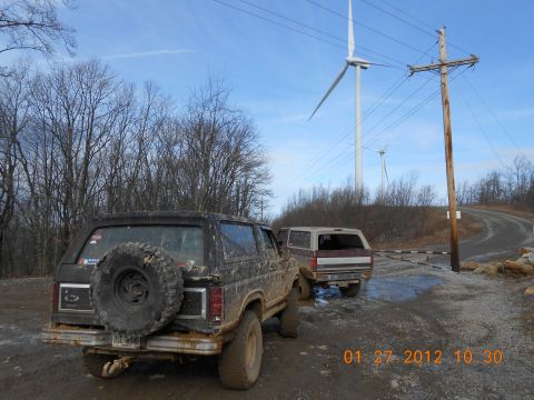 1986 Ford Bronco - Two off-road vehicles are parked on a muddy path with wind turbines in the distance.