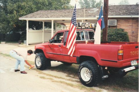 1989 Ford Bronco - A red Toyota pickup truck, modified with a lift kit and large tires, flies American and Christian flags.