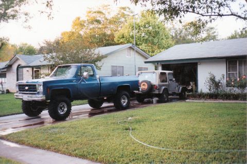 1989 Ford Bronco - A blue pickup truck tows a Jeep Wrangler past a residential garage.