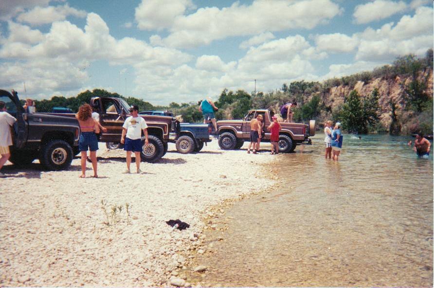 Several trucks parked on a rocky shore next to a shallow river with people around