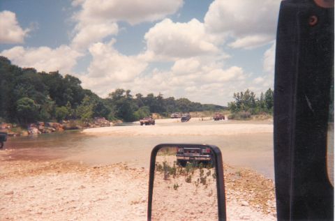 1989 Ford Bronco - Several off-road vehicles navigate a shallow riverbed under a cloudy sky.