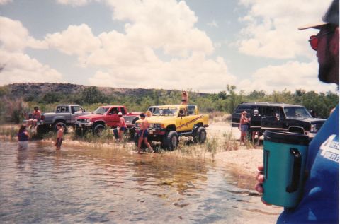 1989 Ford Bronco - A group of off-road vehicles and people gathered by the water on a sunny day.