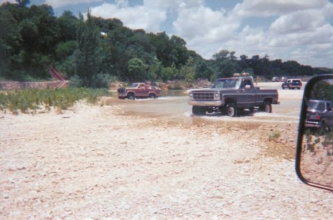 1989 Ford Bronco - Multiple trucks navigate a water crossing on a sunny day with scattered clouds.