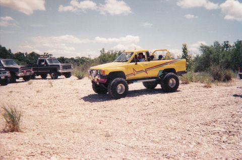 1989 Ford Bronco - A lifted yellow pickup truck with custom graphics sits in a dry, rocky area alongside other vehicles.