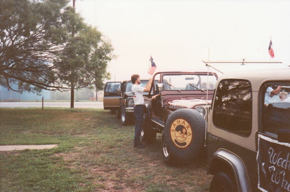 Line of SUVs and a Jeep parked on grass, with a man holding a flag