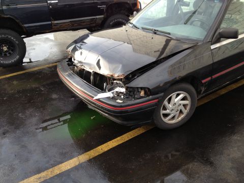 1988 Ford Bronco - A black sedan with significant front-end damage is parked next to a black SUV in a wet parking lot.