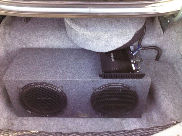 Car trunk interior with a dual subwoofer box and a black amplifier. The spare tire cover is partially open