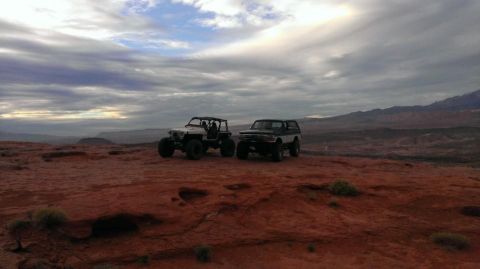1994 Ford Bronco - Two off-road vehicles are parked on a red rock formation with mountains in the background under a dramatic sky.