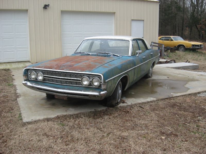 1968 Ford Torino with rusty hood and peeling paint, parked in front of a garage. Yellow car in background