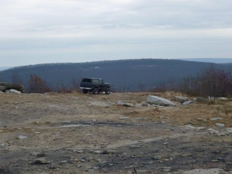 1995 Ford Bronco - A black 1995 Ford Bronco sits on a rocky overlook, with a vast, tree-covered mountain range stretching out behind it under a cloudy sky.
