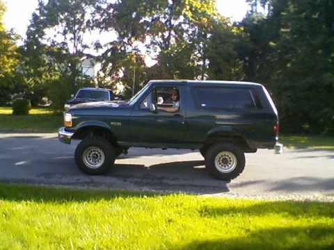 1995 Ford Bronco - A green Ford Bronco is driven on a paved road, with a dark SUV parked in the background.
