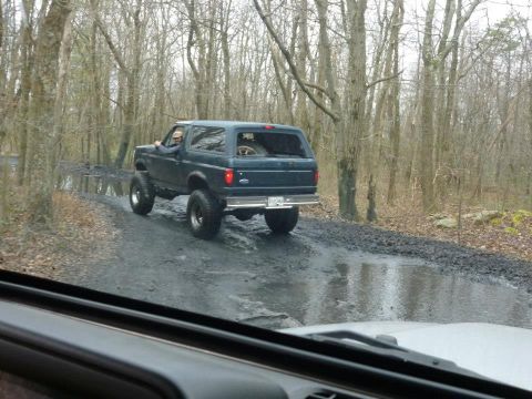 1995 Ford Bronco - A lifted Ford Bronco navigates a wet, wooded path, kicking up mud as it goes.