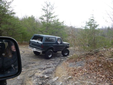 1995 Ford Bronco - A lifted Ford Bronco navigates a rocky, wooded trail, with another vehicle's side mirror visible in the foreground.
