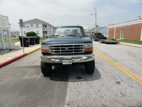 1995 Ford Bronco - A dark teal Ford Bronco is parked on a street with a trailer and another truck visible in the background. The Bronco is facing the camera w…