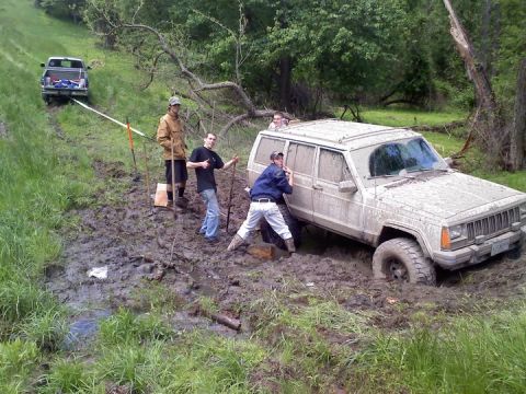 1995 Ford Bronco - A group of people are attempting to pull a mud-covered Jeep Cherokee out of a muddy ditch using a tow strap attached to a pickup truck.