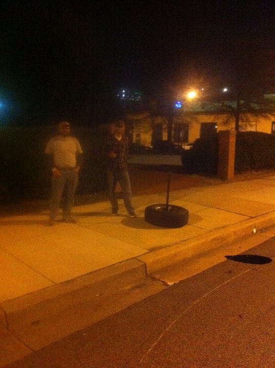 Two men stand on a sidewalk at night next to a tire with a pole through it. A building and streetlights are in the background