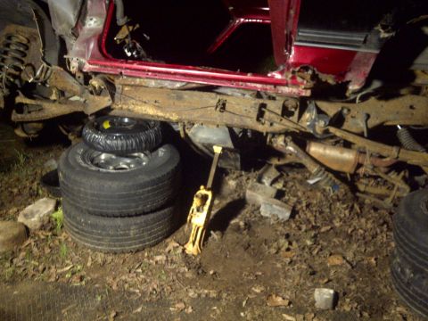 1995 Ford Bronco - The damaged chassis of a red SUV is supported by stacked tires and a jack, revealing extensive rust and mechanical components.
