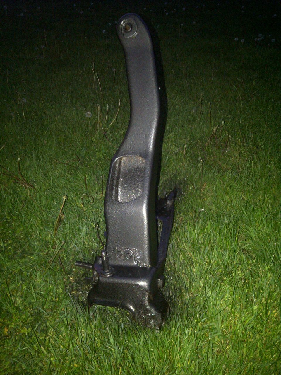 Black metal automotive part, possibly a suspension component, resting on grass. Has some stamped numbers