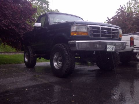 1995 Ford Bronco - A dark blue Ford F-Series truck is parked on a wet asphalt surface next to a white truck.