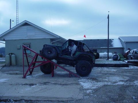 1992 Ford Bronco - Modified off-road vehicle with large tires climbing a red metal ramp in a snowy outdoor area.
