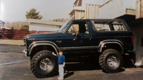 1995 Ford Bronco - A child stands next to a lifted dark blue 1995 Ford Bronco, its large tires and classic SUV profile.