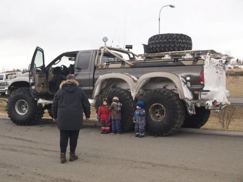 1978 Ford Bronco - A heavily modified pickup truck with six wheels and massive tires is parked on a street.