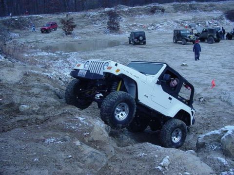 1992 Ford Bronco - A white Jeep Wrangler Rubicon navigates a steep, rocky incline during an off-road event, with other vehicles and people in the background.