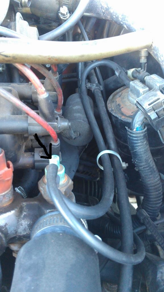 Close-up of engine bay with various hoses and electrical connectors. An arrow points to a sensor