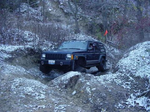1992 Ford Bronco - A dark blue Jeep Cherokee XJ navigates a snowy, wooded trail with a red flag marking the path.