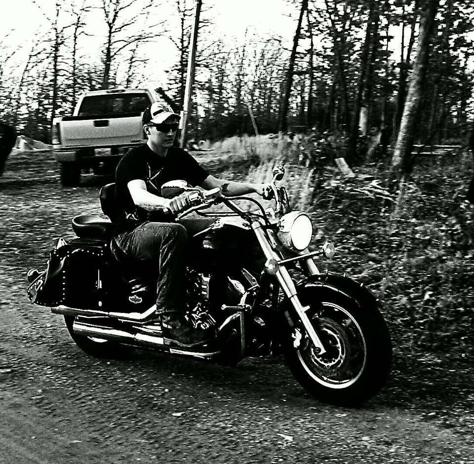 Man riding a cruiser motorcycle on a dirt road, with a pickup truck behind him in a wooded area