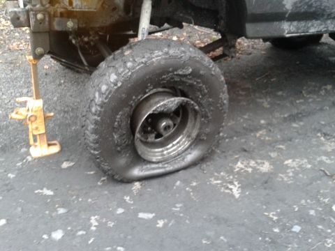 1995 Ford Bronco - A flat tire on a vehicle is shown, supported by a yellow jack. The tire and surrounding ground are covered in mud.