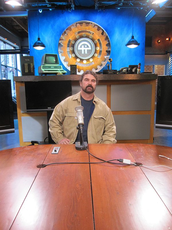 Man in tan jacket sits at a wooden table with a microphone in front of a blue backdrop with a large gear graphic