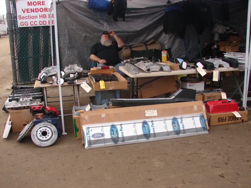 Vendor sitting behind tables piled with automotive parts, including Ford service parts boxes and engine components
