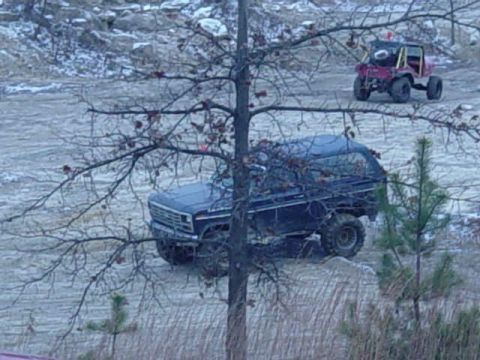1992 Ford Bronco - A dark blue SUV navigates a muddy terrain, with another off-road vehicle visible in the distance.