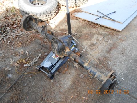 1986 Ford Bronco - A vehicle's rear axle assembly is shown supported by a jack on the ground, with a spare tire and tools in the background.