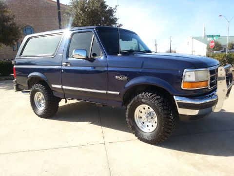 1995 Ford Bronco - This blue Ford Bronco is shown from the front three-quarter view, parked on a paved surface under a clear sky.