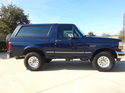 1995 Ford Bronco - A dark blue Ford Bronco SUV is shown from the side, parked on a paved surface under a clear blue sky.