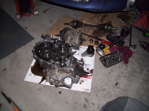 1995 Ford Probe - Transmission and drive axles are laid out on a piece of cardboard on the garage floor, surrounded by tools and parts.
