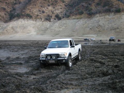 1985 Ford Bronco - A white pickup truck navigates through a muddy terrain with other vehicles in the background.