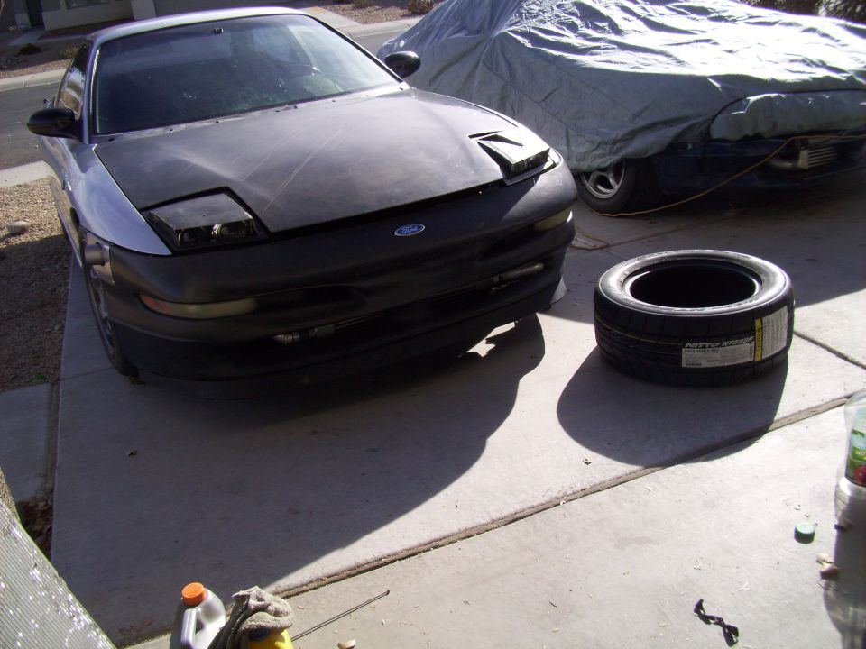 Front view of a 1995 Ford Probe with a black hood and a tire next to it. Another car is covered in a tarp behind it