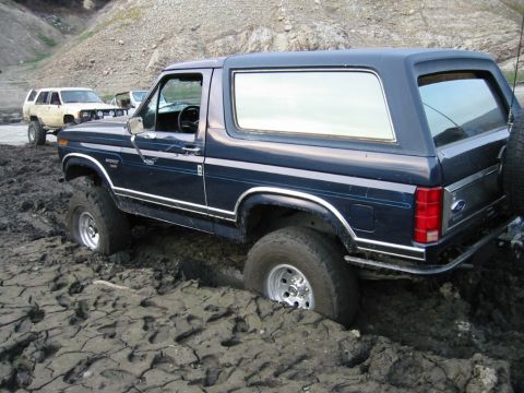 1985 Ford Bronco - A dark blue 1985 Ford Bronco is stuck in thick mud, with another SUV visible in the background.