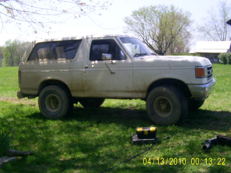 White Ford Bronco parked on grass, side view. Mud splatters on body. Aftermarket wheels. Toolbox on ground