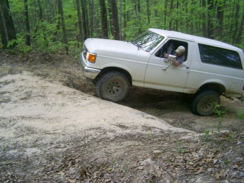1990 Ford Bronco - A white 1990 Ford Bronco navigates a challenging off-road trail through a wooded area.