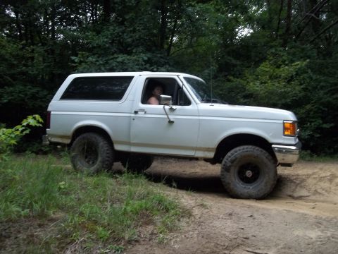 1990 Ford Bronco - A white 1990 Ford Bronco is driven off-road through a wooded area on a dirt path.