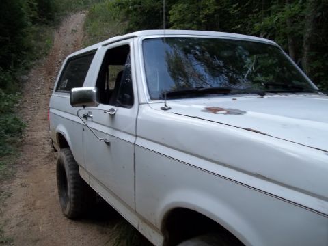 1990 Ford Bronco - White Ford Bronco drives on a dirt road through a wooded area.