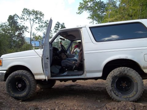1990 Ford Bronco - A white 1990 Ford Bronco is seen from the side with the driver's door ajar, revealing the interior and a person inside.