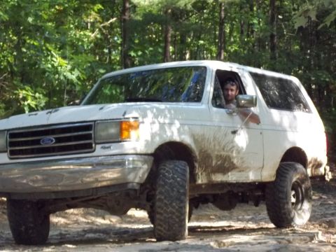 1990 Ford Bronco - A white 1990 Ford Bronco is shown driving off-road through a wooded area, with mud splattered on its body.